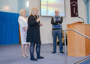 Lucy is wearing a black dress and the chains of office as she speaks into a microphone. Her consort is wearing a white dress behind her as Jeff watches from the podium. In the background a sign reads Thank you Jesus.