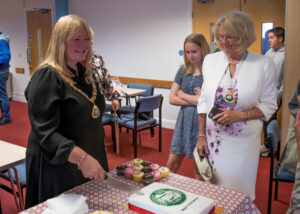 Lucy smiles as she cuts into a cake decorated with the Peacehaven Town Council logo as her consort and guests watch.